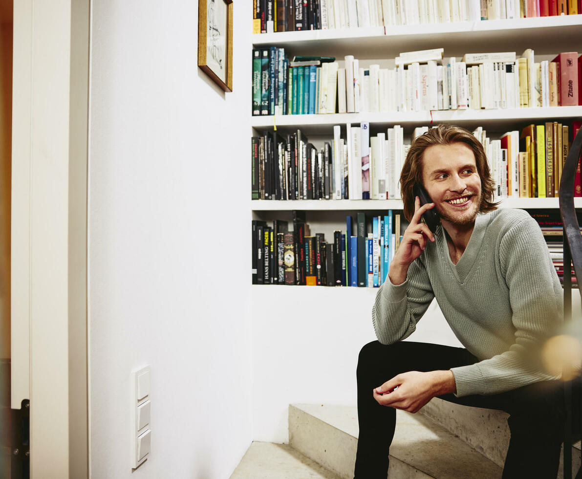 Smiling male sitting on stairs, having a phonecall. bookshelf in background
 Smiling male sitting on stairs, having a phonecall. bookshelf in background